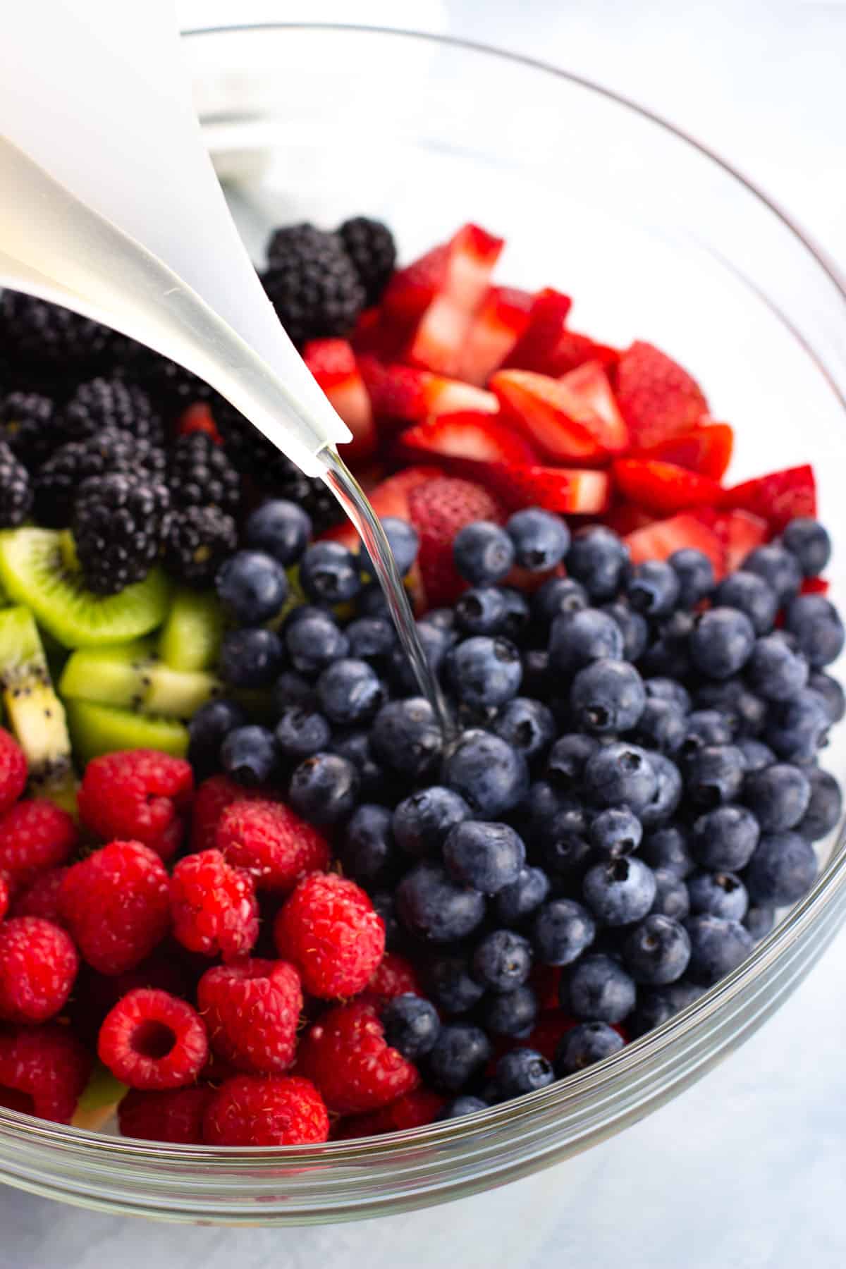 Simple syrup being poured into a bowl of sliced fruit and berries.