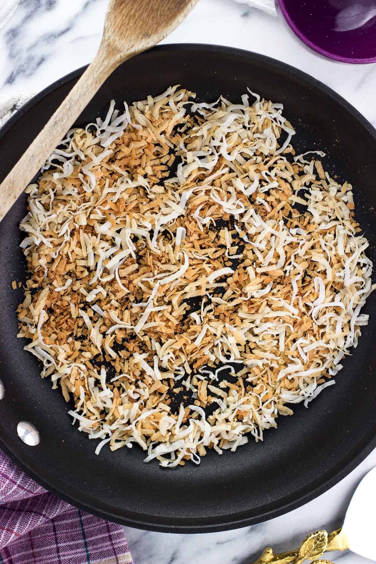 Toasted coconut in a skillet with a wooden spoon.