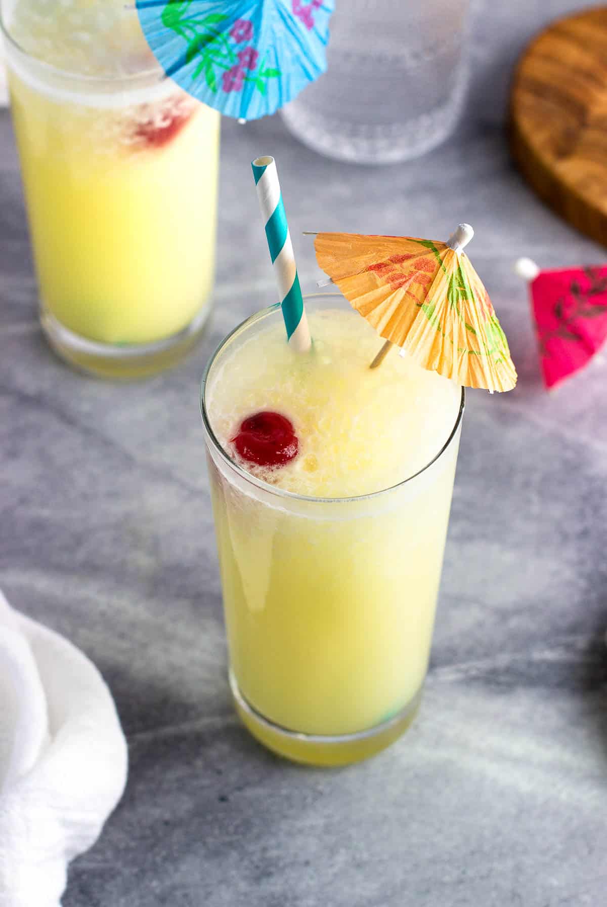 A pina colada mocktail in a glass with a straw, paper drink umbrella, and maraschino cherry.