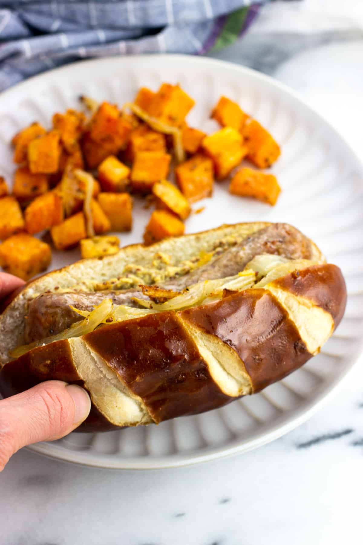 Bratwurst in a pretzel bun on a plate with sweet potatoes.