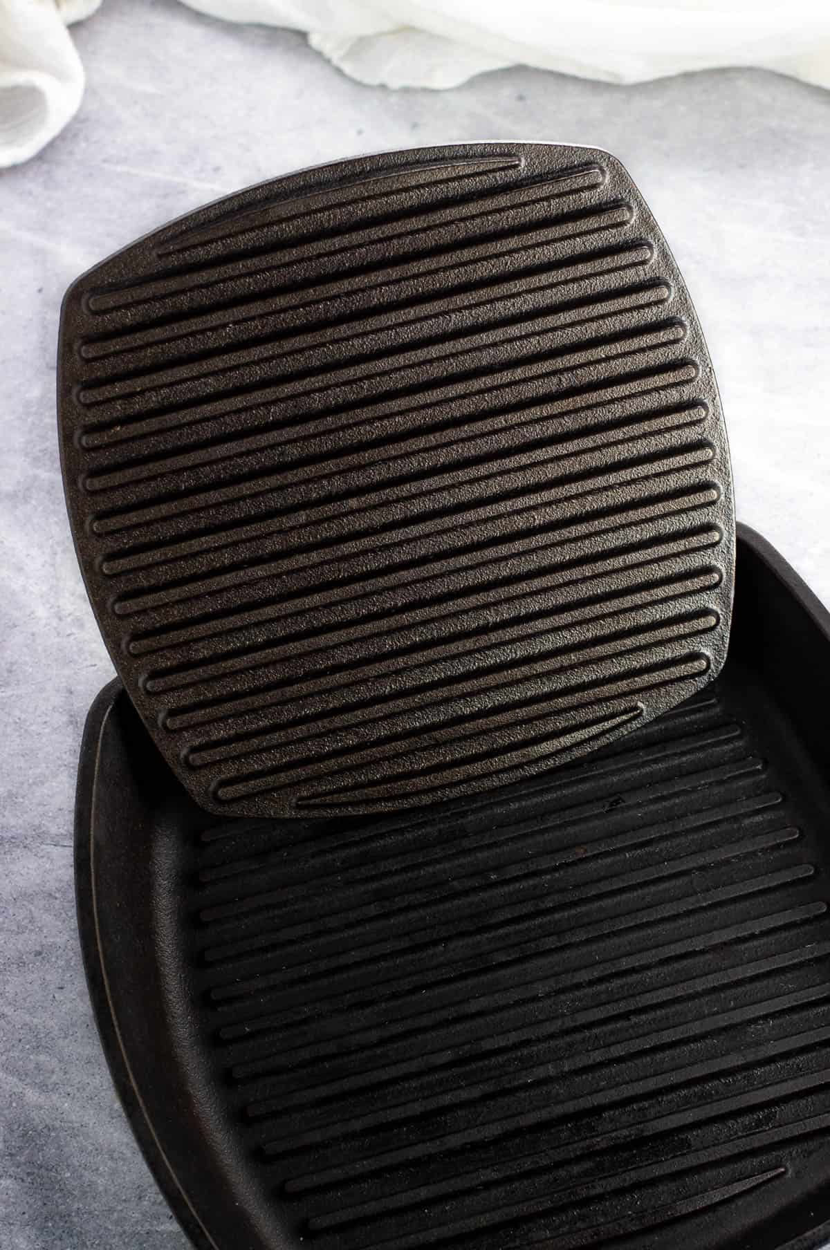 A cast iron panini press propped up inside a grill pan showing the grill surfaces.