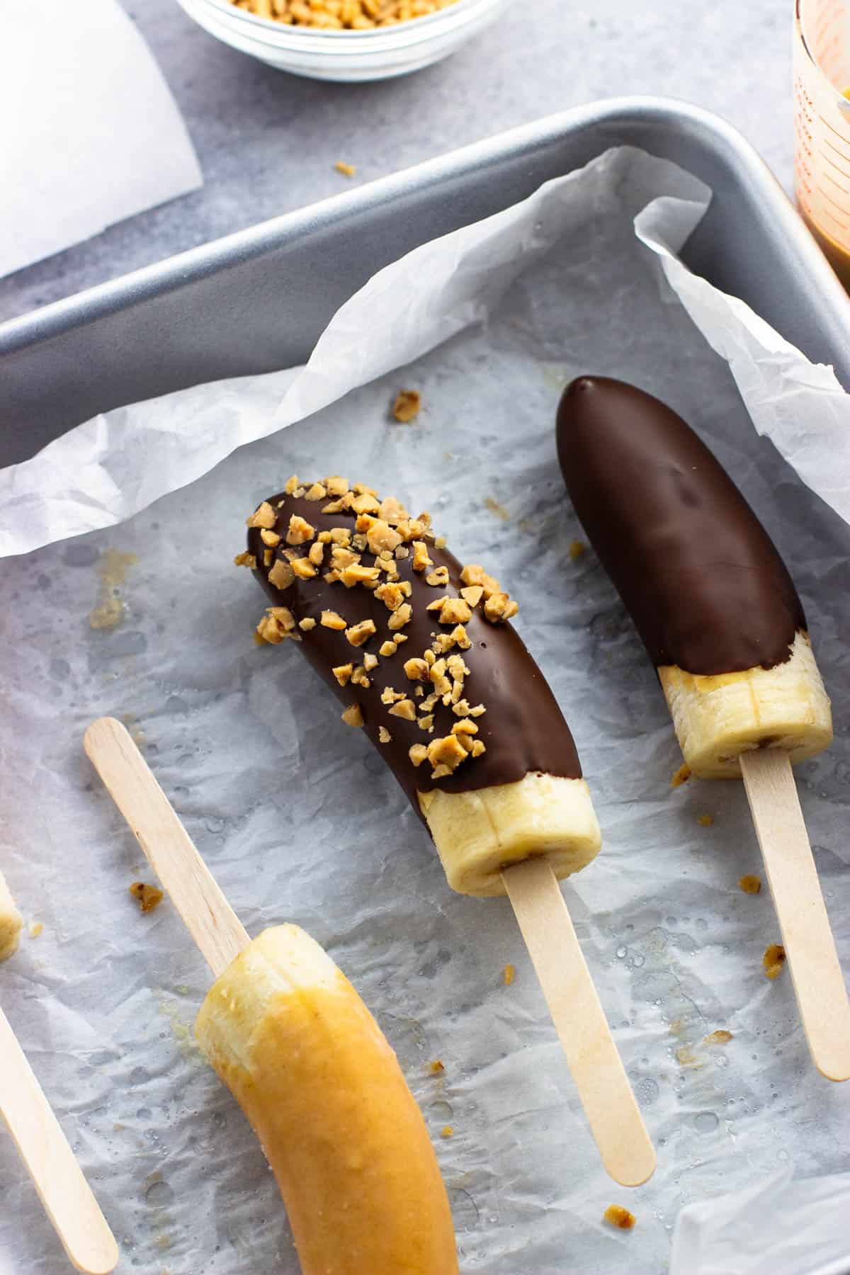 Frozen chocolate banana popsicles in a dish with one coated in chopped peanuts.