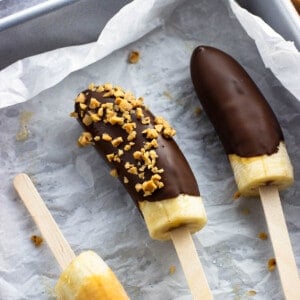 Frozen chocolate banana popsicles in a dish with one coated in chopped peanuts.