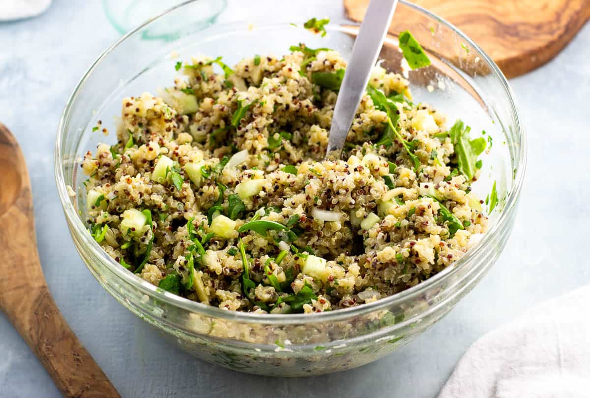 A large glass serving bowl filled with herbed quinoa salad with a metal spoon in the center.