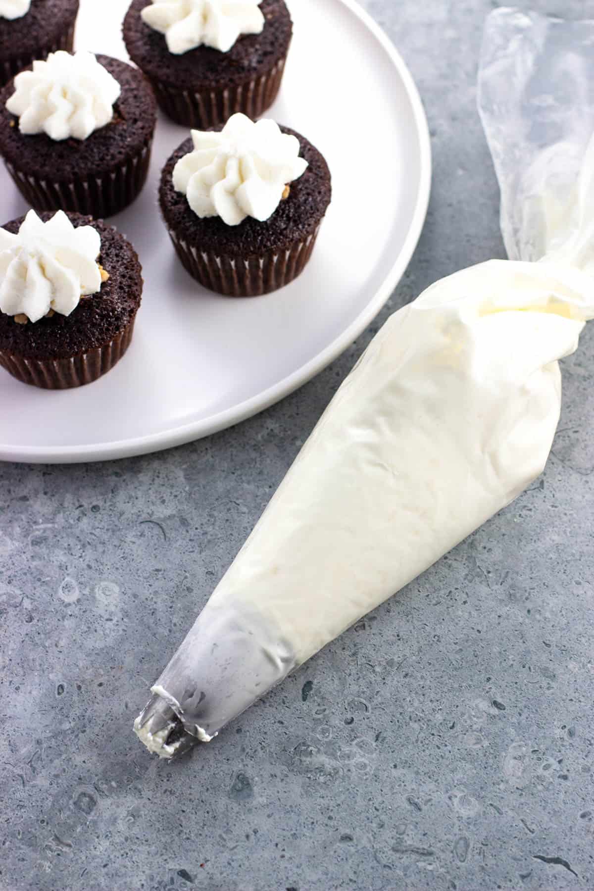 A full piping bag of whipped cream next to chocolate cupcakes dolloped with whipped cream.