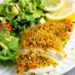 A panko baked cod fillet cut in half on a plate with salad and lemon wedges.