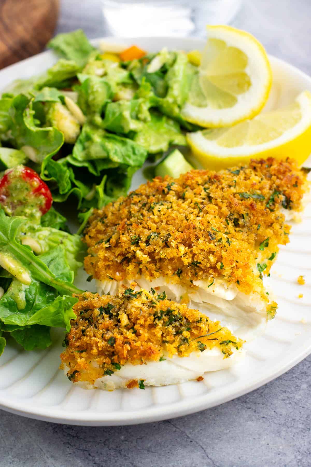 A panko baked cod fillet cut in half on a plate with salad and lemon wedges.