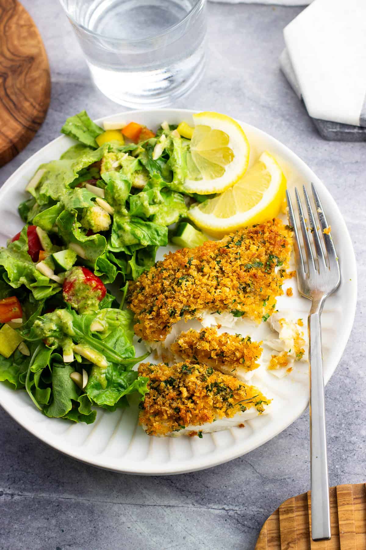 A panko crusted cod fillet on a dinner plate with a side salad and lemon wedges.