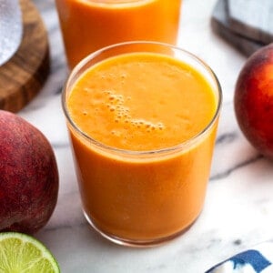 A peach mango smoothie in a short glass surrounded by fresh peaches and limes.