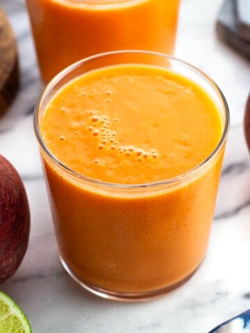 A peach mango smoothie in a short glass surrounded by fresh peaches and limes.