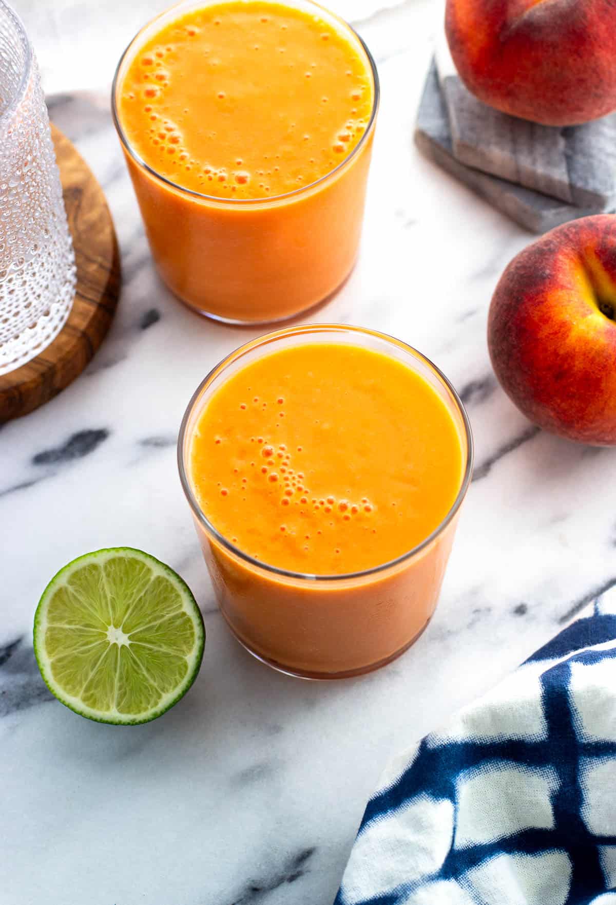 Two glasses of smoothie on a marble board next to peaches and limes.