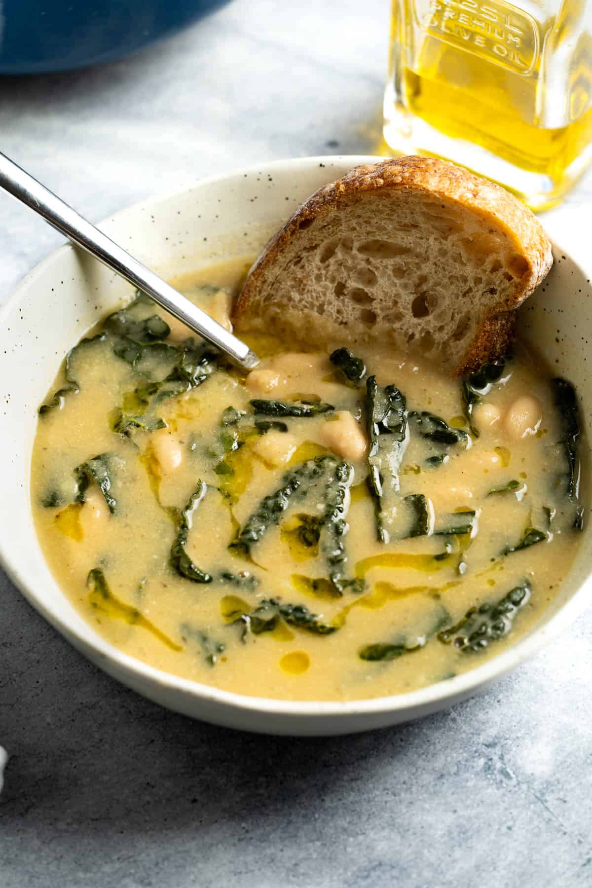 A bowl of potato and white bean soup drizzled with olive oil with a piece of bread stuck in.