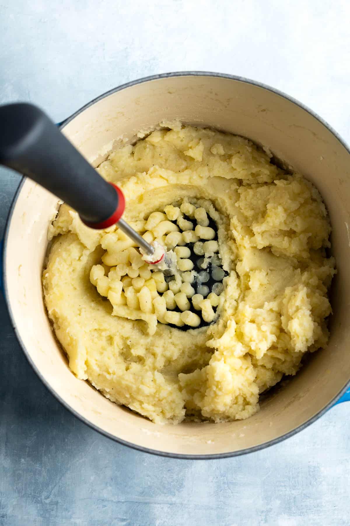 The mashed potatoes being combined with a handheld masher in a pot.
