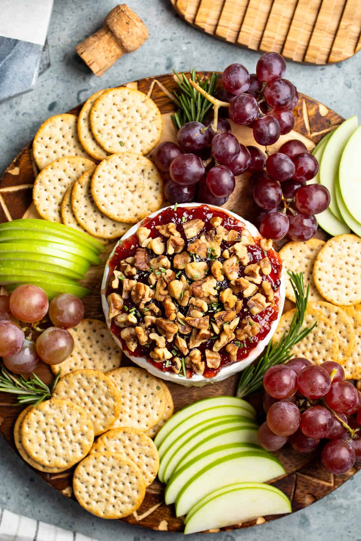 Jam and walnut-topped baked brie on a board with crackers, apple slices, and grapes.