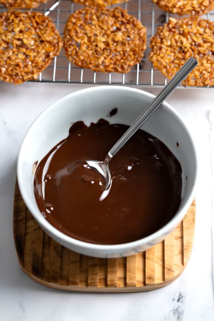 A bowl of melted chocolate in front of florentines on a wire rack.