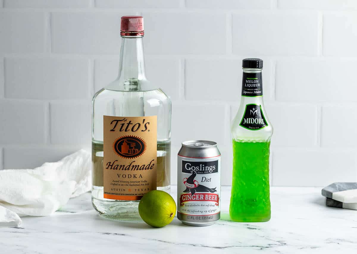 Vodka, ginger beer, Midori, and a lime on a countertop.