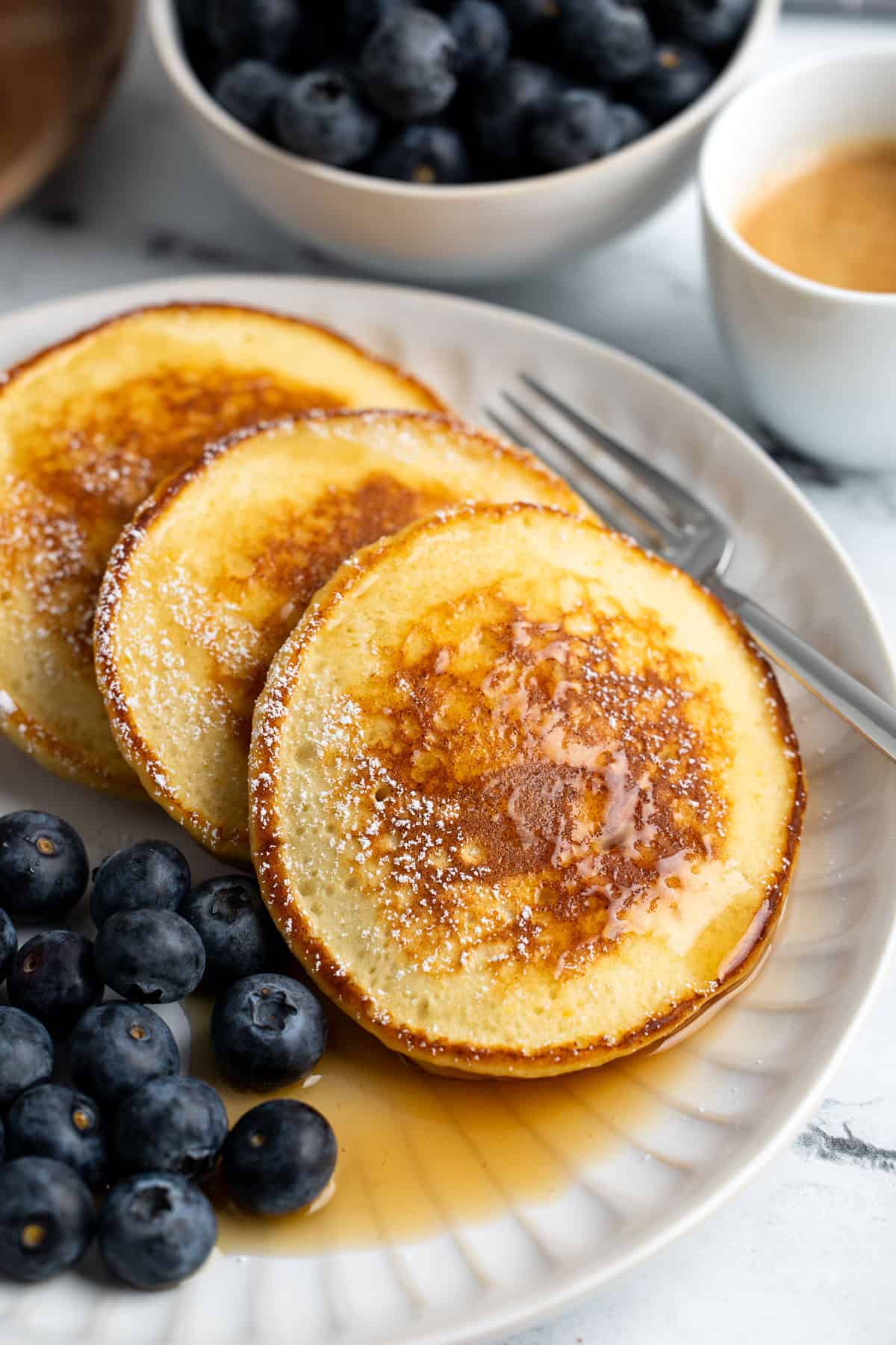 Three maple syrup-drizzled ricotta pancakes on a plate with fresh blueberries.