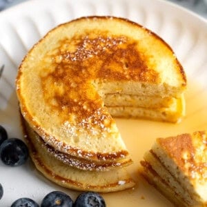 A stack of three ricotta pancakes on a plate with a wedge cut out all drizzled with maple syrup.