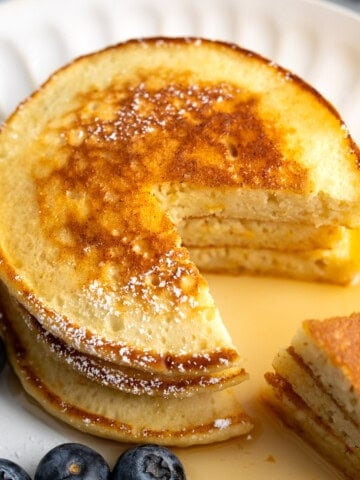 A stack of three ricotta pancakes on a plate with a wedge cut out all drizzled with maple syrup.