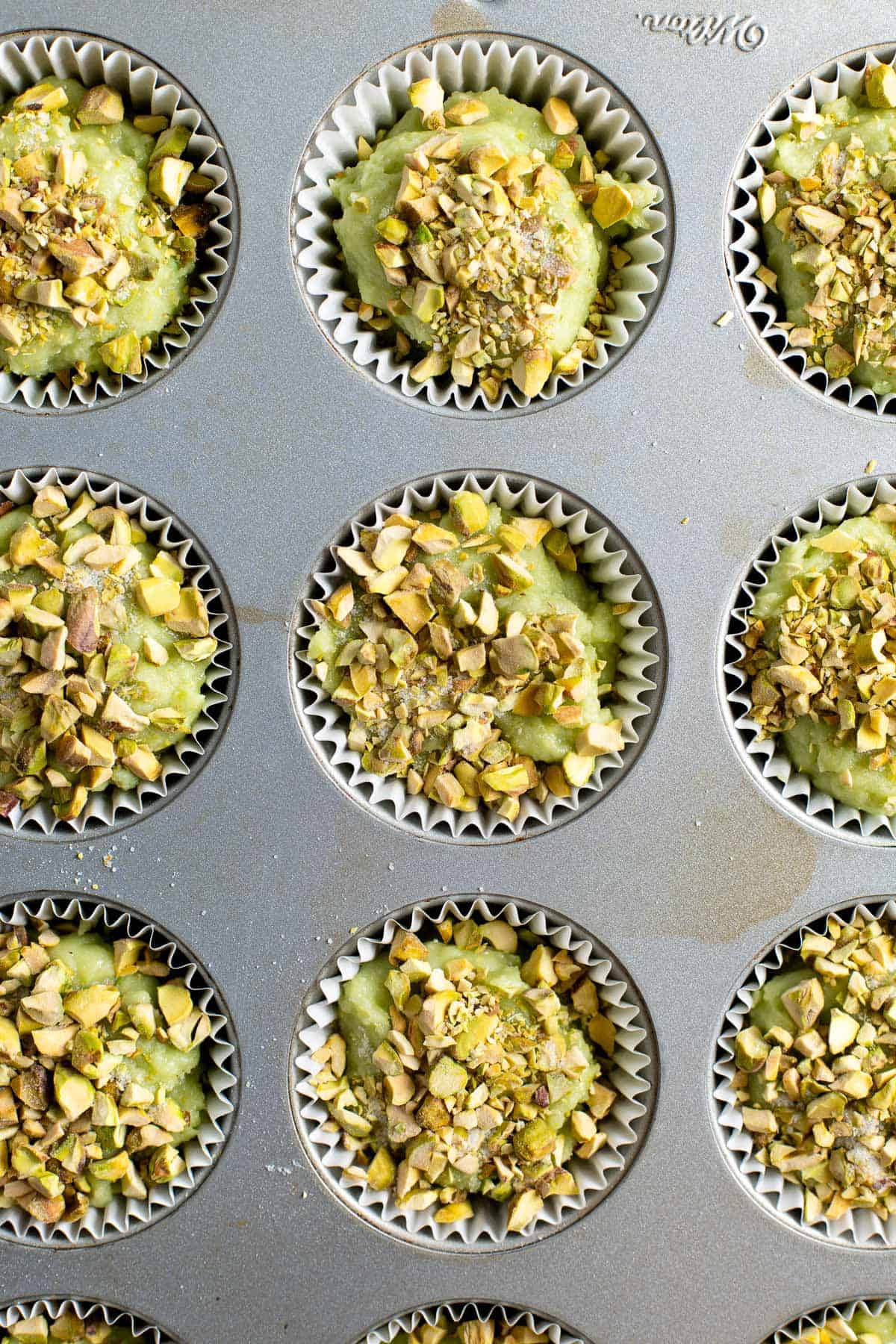 Muffin batter dolloped into the muffin tin and topped with chopped pistachios.
