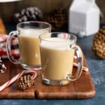 Two peppermint tea lattes in glass mugs on a board next to pinecones and candy canes.