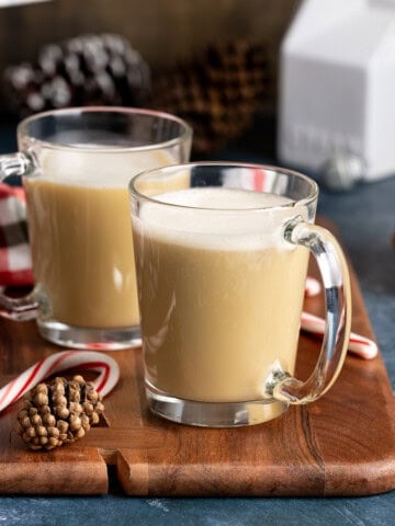 Two peppermint tea lattes in glass mugs on a board next to pinecones and candy canes.