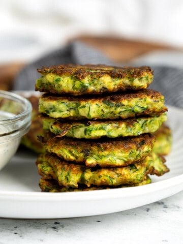 A stack of pan fried Italian-style zucchini fritters on a plate next to a bowl of dipping sauce.