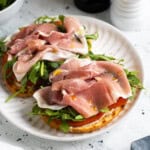 An open-face prosciutto sandwich on a plate with lemon aioli, tomato slices, arugula, prosciutto, olive oil, salt, and pepper.