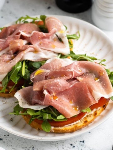 An open-face prosciutto sandwich on a plate with lemon aioli, tomato slices, arugula, prosciutto, olive oil, salt, and pepper.