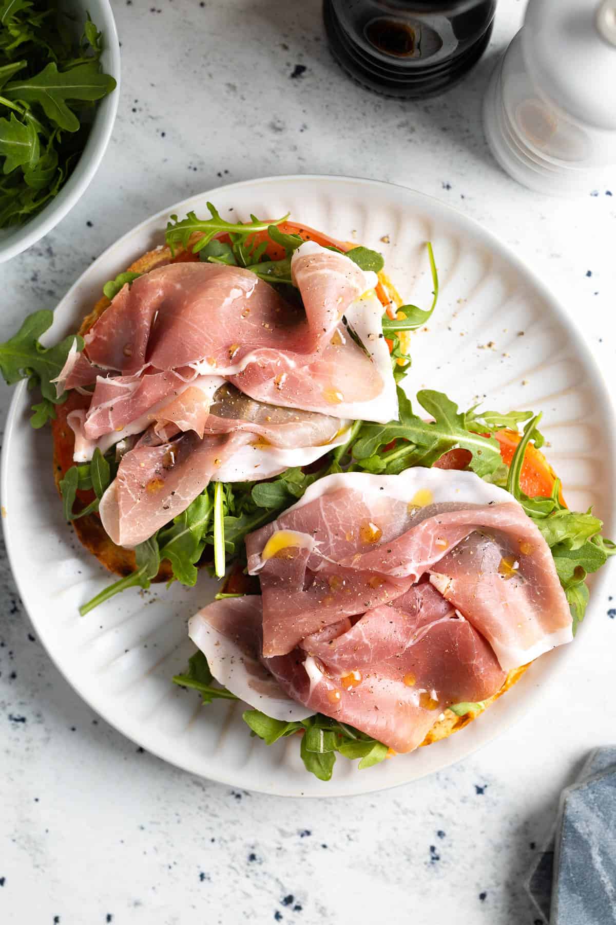 Two slices of bread topped with lemon aioli, tomato, arugula, and prosciutto slices.