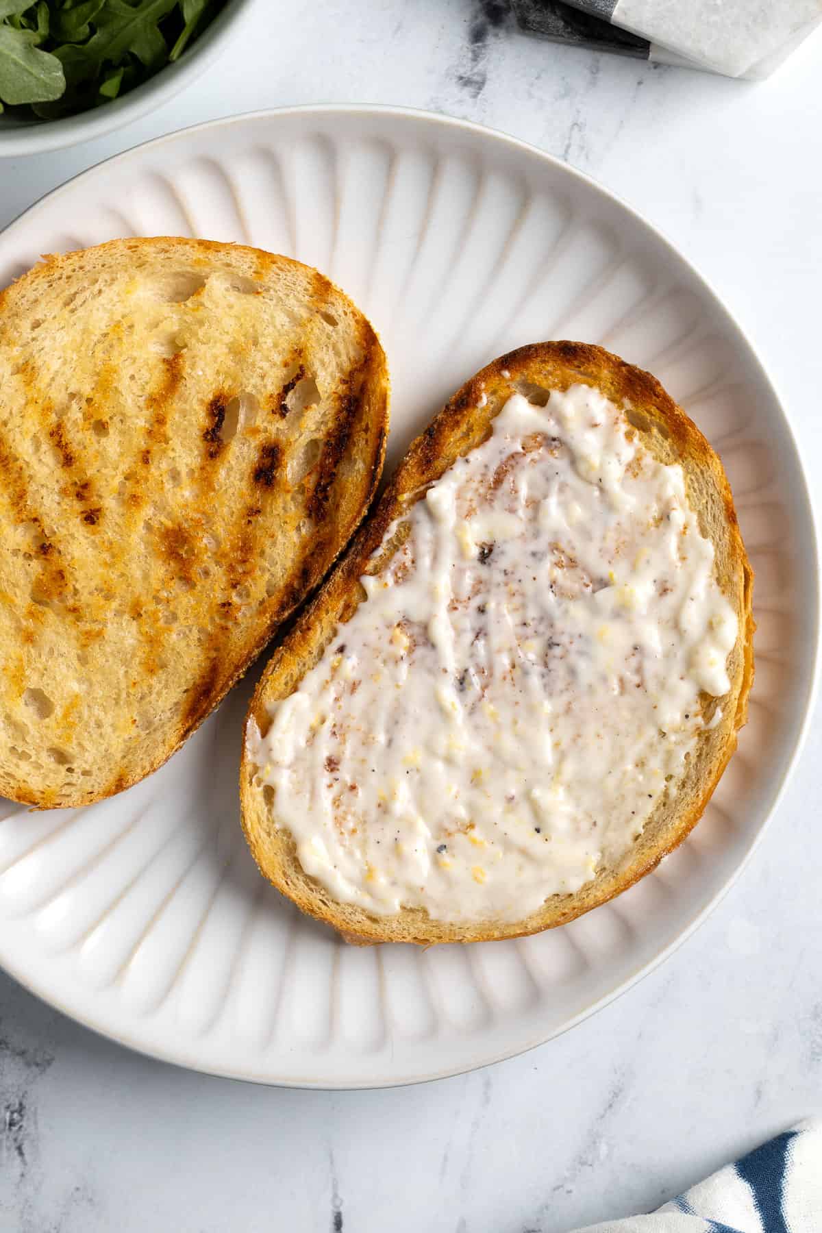 Lemon aioli spread onto one of two slices of grilled bread on a plate.