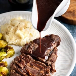 Red wine jus being poured over a half-sliced steak on a plate with mashed potatoes and brussels sprouts.