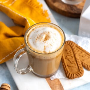 A cinnamon-dusted cookie butter latte in a glass mug next to two Biscoff cookies.