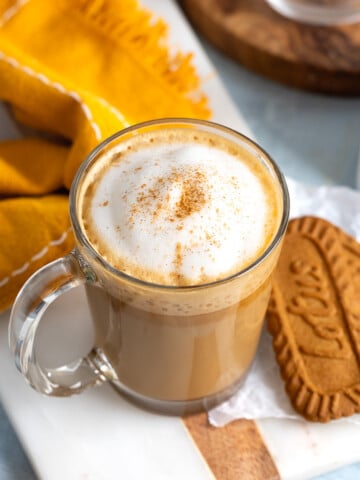 A cinnamon-dusted cookie butter latte in a glass mug next to two Biscoff cookies.