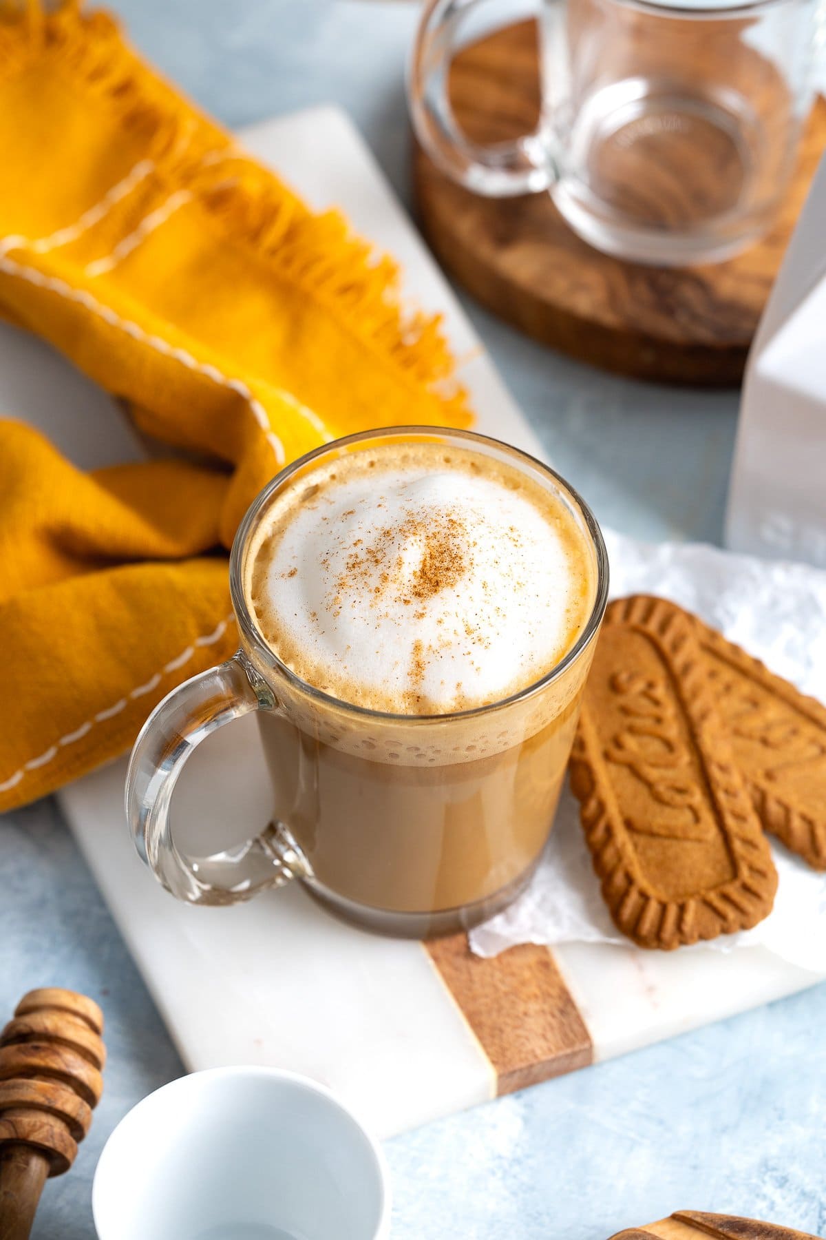 A cinnamon-dusted cookie butter latte in a glass mug next to two Biscoff cookies.