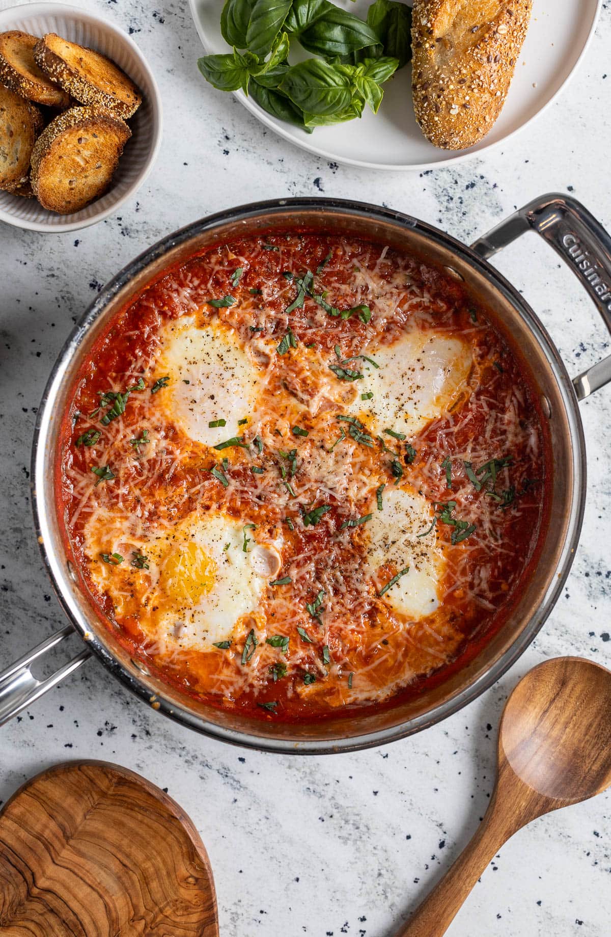 Eggs in purgatory topped with shredded Parmesan and fresh basil in a pan next to crusty bread.