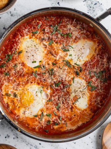 Eggs in purgatory topped with shredded Parmesan and fresh basil in a pan next to crusty bread.