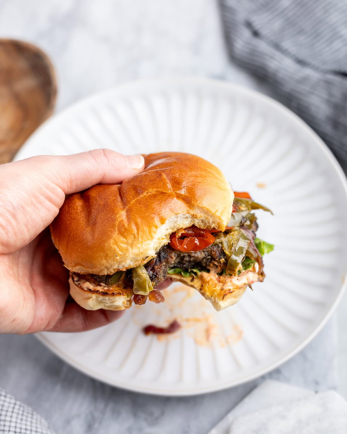 A hand holding an Italian sausage burger topped with aioli and peppers and onions.