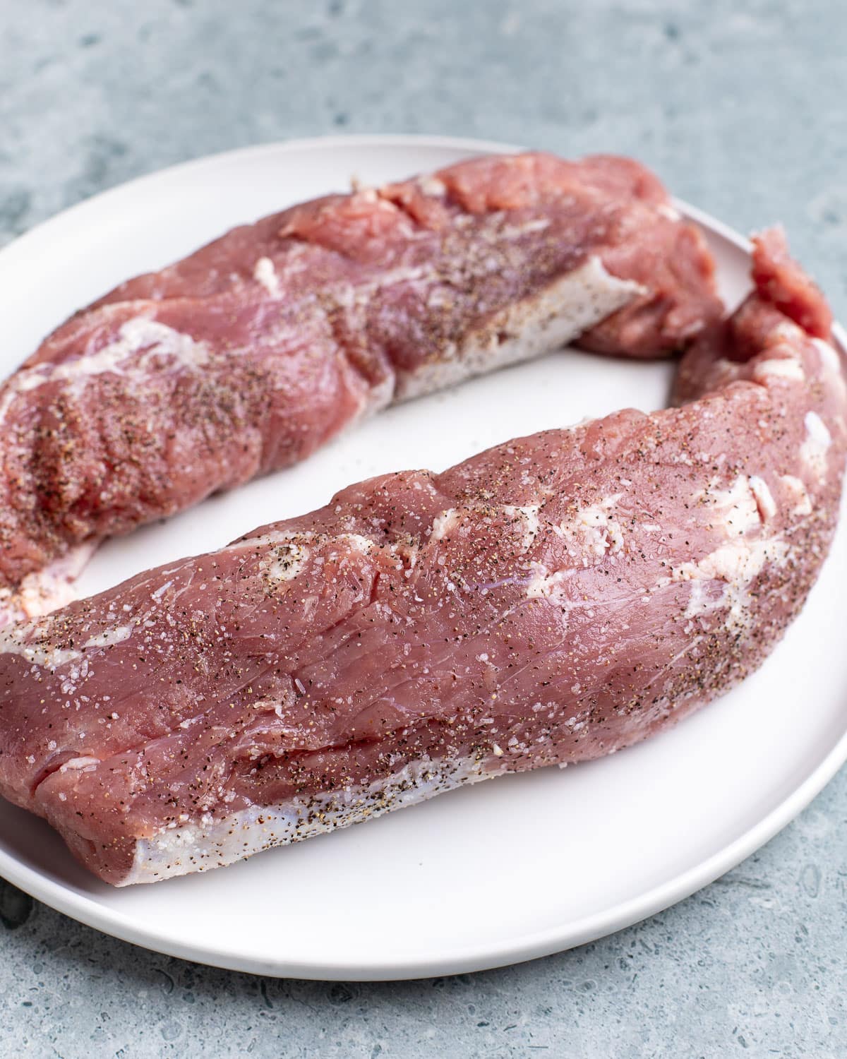 Raw pork tenderloin segments on a plate coated in salt and pepper.