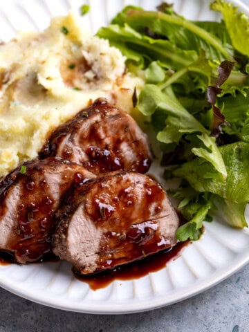A plate of sliced pork tenderloin with raspberry sauce, salad, and mashed potatoes.