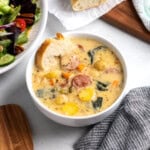 A bowl of sausage, potato, and white bean soup with a slice of baguette dunked into it.