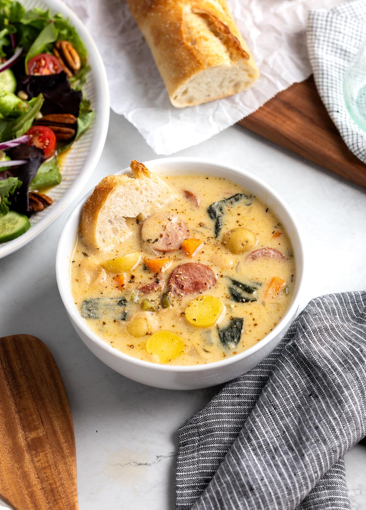 A bowl of sausage, potato, and white bean soup with a slice of baguette dunked into it.
