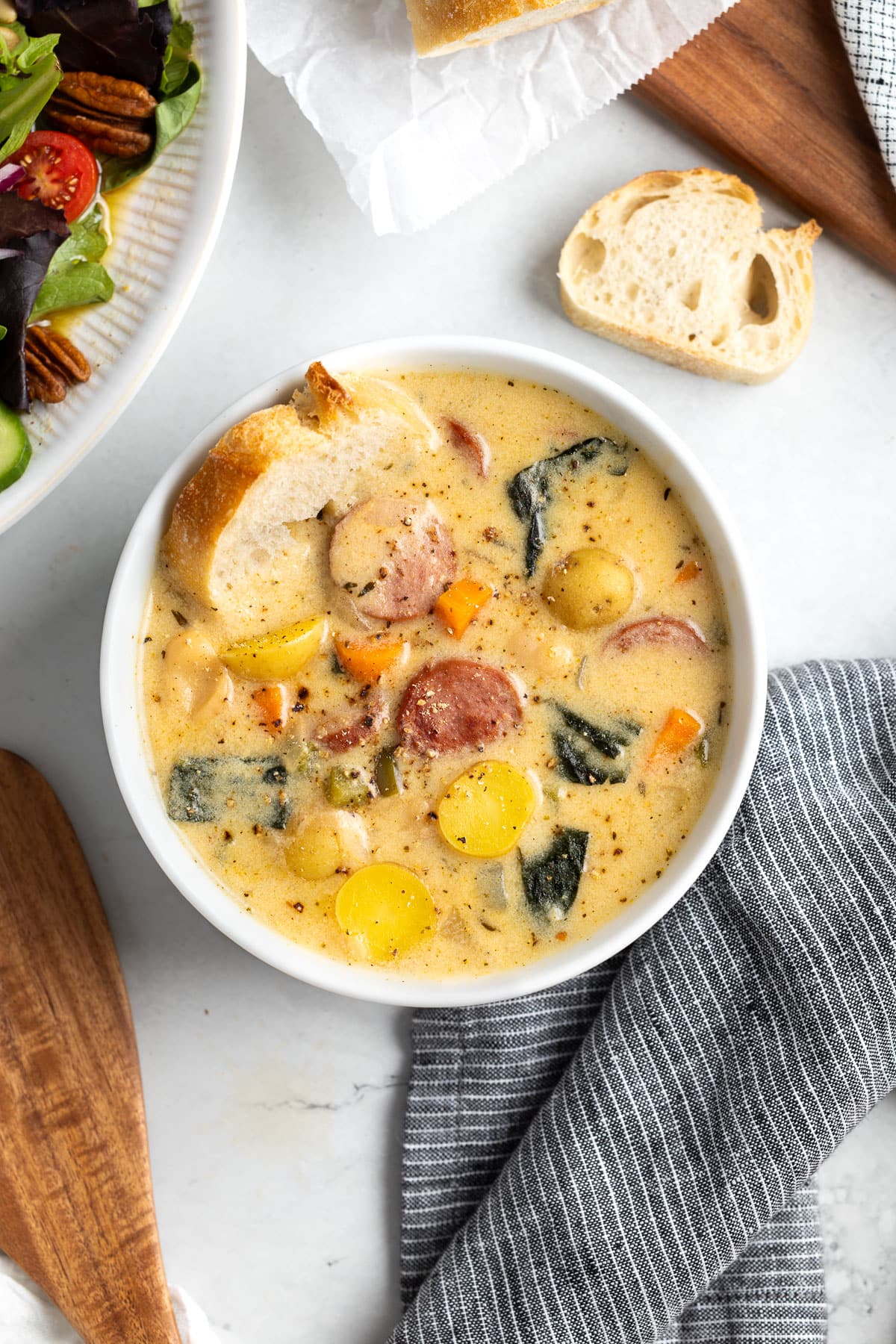 A bowl of sausage, potato, and white bean soup with a slice of baguette dunked into it.