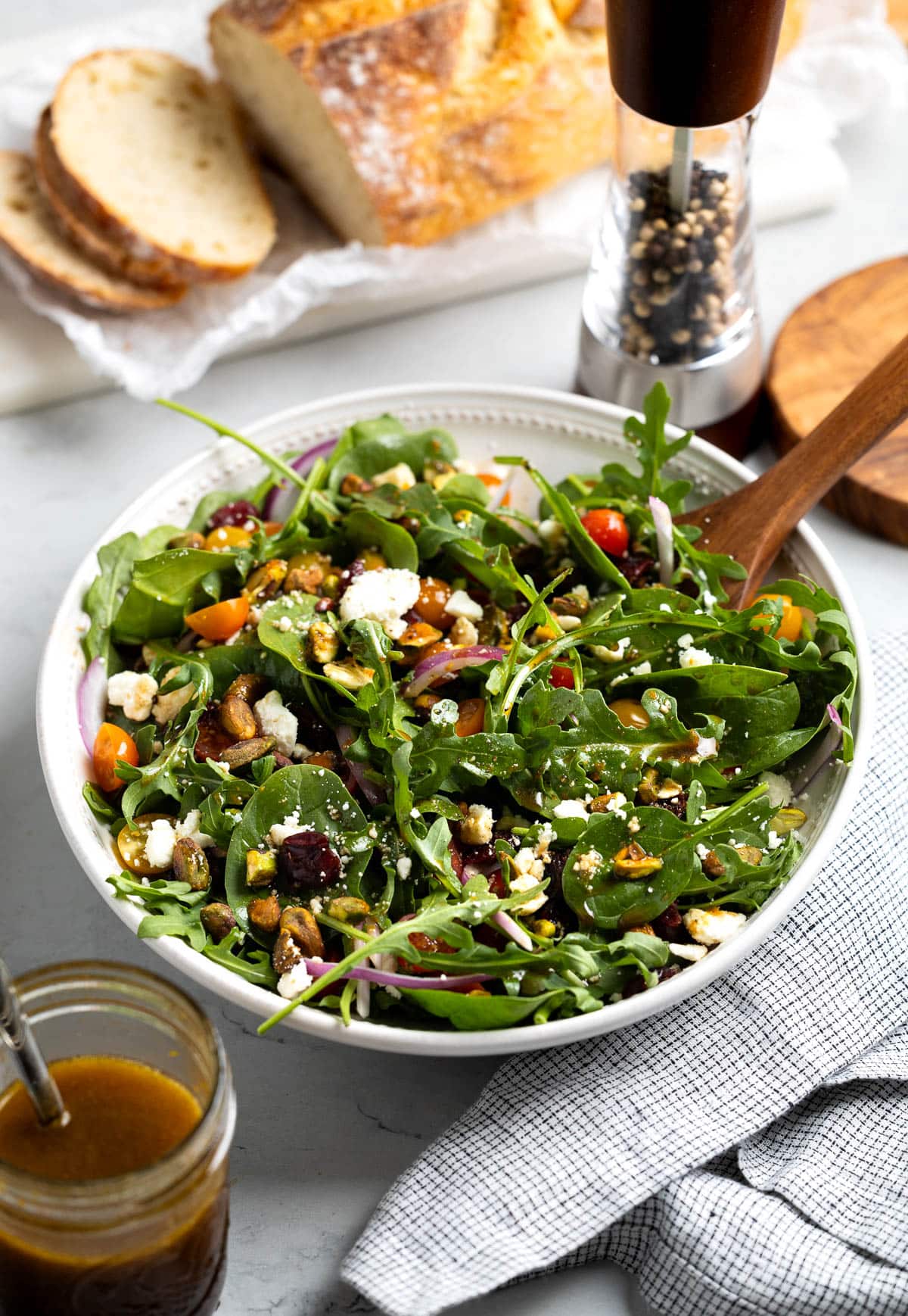 A bowl of arugula and spinach salad next to a sliced loaf of bread and a pepper mill.
