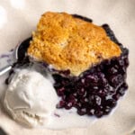 A luscious blueberry cobbler serving on a scalloped plate with vanilla ice cream.