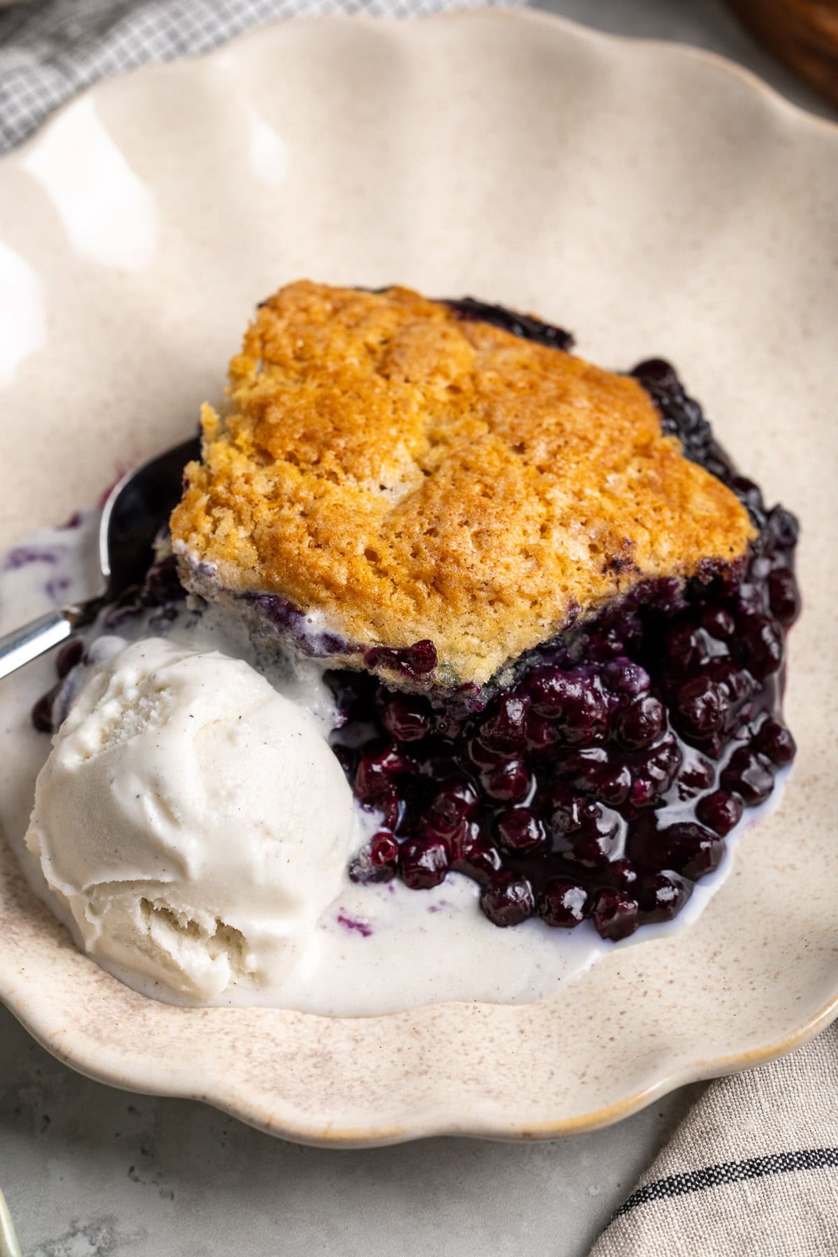 A luscious blueberry cobbler serving on a scalloped plate with vanilla ice cream.