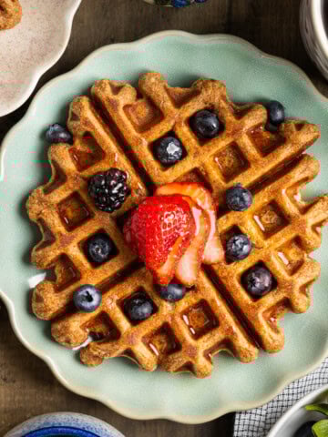 Whole wheat buttermilk waffles on plates topped by fresh berries and maple syrup.