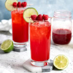 Two drinks in highball glasses garnished with raspberries and lime rounds.