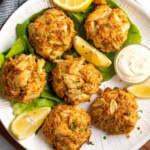 A plate of six crab cakes with lemon wedges and a bowl of aioli.