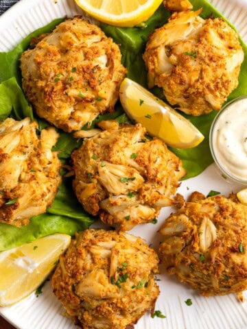 A plate of six crab cakes with lemon wedges and a bowl of aioli.